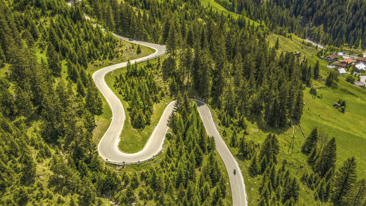 Luftaufnahme einer Bergstraße mit vier Kehren durch Fichtenwald, ein einzelnes Motorrad, rechts die Häuser eines Dorfs Luftaufnahme einer Bergstraße mit vier Kehren durch Fichtenwald, ein einzelnes Motorrad, rechts die Häuser eines Dorfs