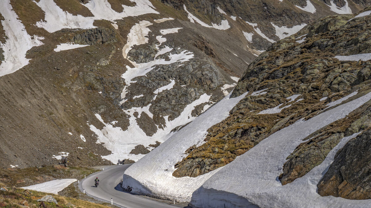 Zwei Motorräder in einer Haarnadelkurve, seitlich eine hohe Wand aus Altschnee, oben Felshänge mit Schneefeldern Zwei Motorräder in einer Haarnadelkurve, seitlich eine hohe Wand aus Altschnee, oben Felshänge mit Schneefeldern